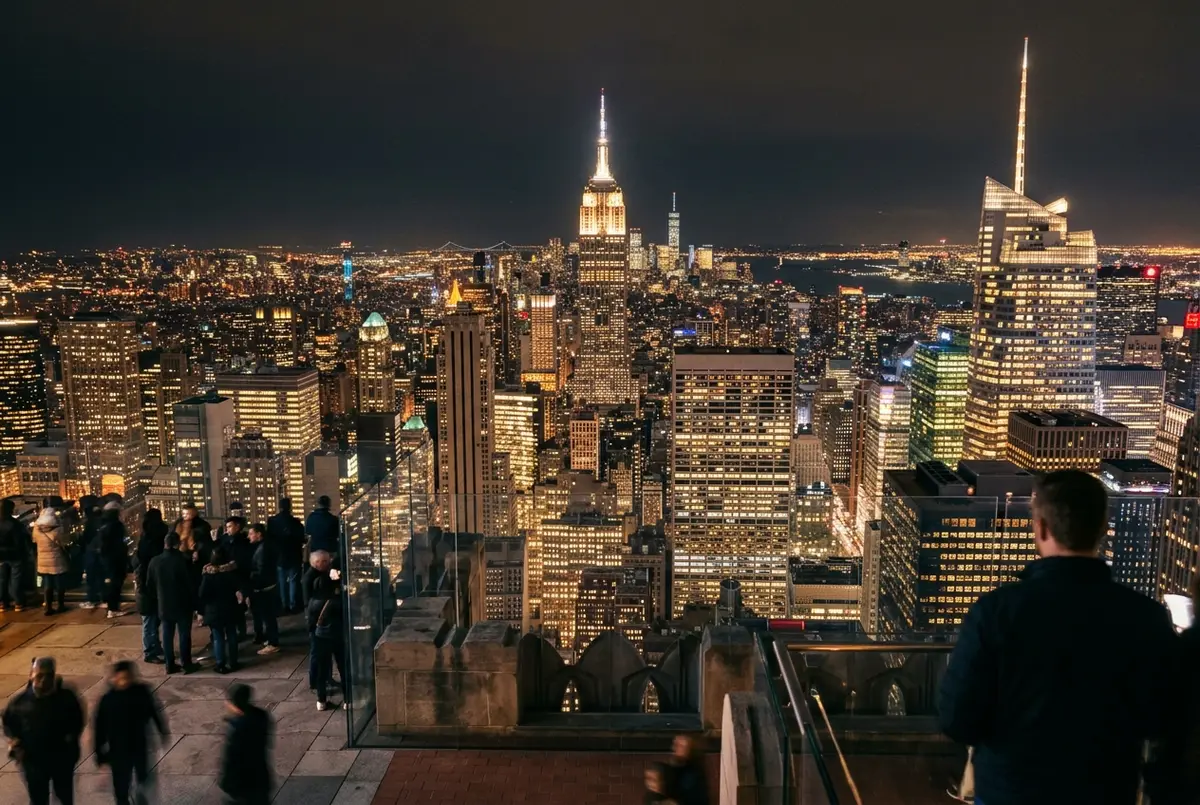 visitantes apoyados en la barandilla del mirador Top of the Rock de noche, perfil de Manhattan iluminado y el Empire State Building brillando al fondo, cielo oscuro